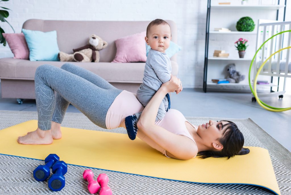 Young happy mother working out on yoga mat with her little child at home