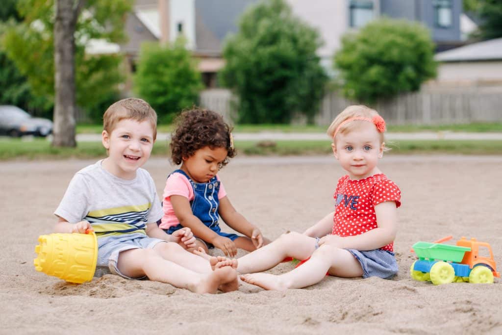 Portrait of three cute Caucasian and hispanic latin toddlers babies children sitting in sandbox playing with plastic colorful toys. Little girls and boy friends having fun together on playground.